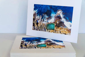 Two prints of a coastal scene with rocks and a greenish-blue object on a white surface.